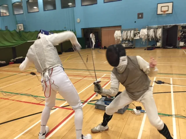 Two foil fencers exchanging attacks in a sports hall